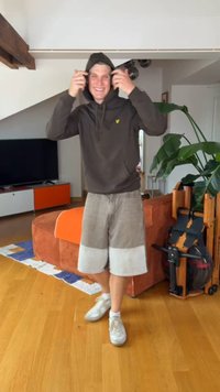 Homme souriant portant un sweat à capuche foncé et un short clair ajuste sa capuche dans un salon avec un sol en bois, une grande plante, des équipements de sport et un canapé orange.