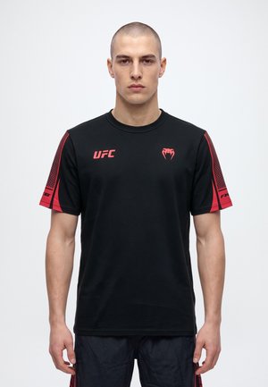 Мъж позира с черна тениска на UFC с червени панели на ръкавите и лога на светъл едноцветен фон.