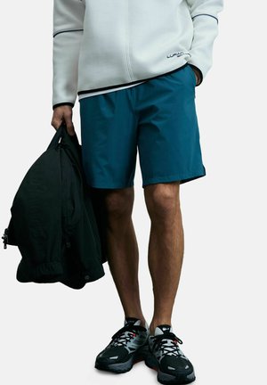 Heren teal korte broek van lichtgewicht stof, gecombineerd met een witte zip-up jas. Houdt een zwarte jas vast. Zwarte en grijze sportschoenen.