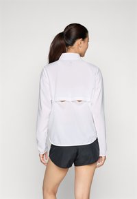 White long-sleeve pullover with a high collar, featuring a layered back design with cutouts. Smooth fabric and a relaxed fit. Black shorts visible.