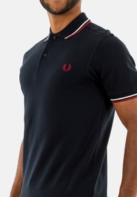 Marinblå polo skjorta i bomull, med en treknapps placket, röd- och vitrandig krage samt en liten broderad logotyp på bröstet.