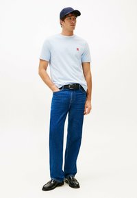 Maglietta di cotone azzurra chiara con un piccolo logo rosso, abbinata a jeans in denim blu. Una cintura nera e scarpe nere lucide completano l'outfit.