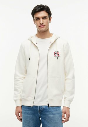 Junger Mann trägt weißen Kapuzenpullover mit Reißverschluss und Tommy Hilfiger-Logo, weißes T-Shirt und blaue Jeans, steht vor einem schlichten hellen Hintergrund.