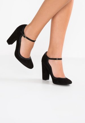 Chaussures à talons hauts en daim noir avec bout rond, bride à la cheville et talon bloc. Texture lisse avec un design classique, présentées en plein mouvement.