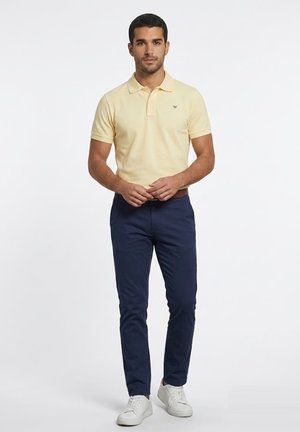 Hombre de pie mirando hacia adelante, vestido con una camisa tipo polo de color amarillo pálido, pantalones azul marino y zapatillas blancas sobre un fondo blanco liso.