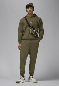 Giovane uomo che indossa un felpa olive green della Jordan, pantaloni jogger abbinati e un berretto, con una borsa a tracolla in camouflage e sneakers bianche e rosse, in piedi.