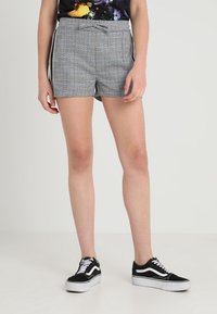 Jambes portant un short à carreaux gris avec des bandes latérales et des baskets noir et blanc, se tenant devant un fond uni.