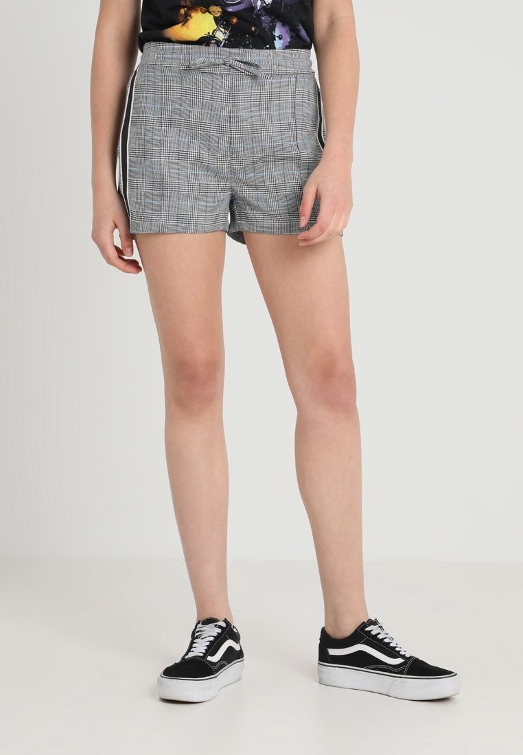Jambes portant un short à carreaux gris avec des bandes latérales et des baskets noir et blanc, se tenant devant un fond uni.