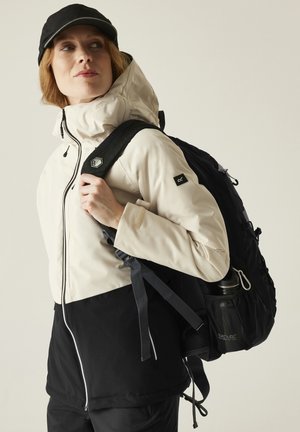 Mujer con gorra negra y chaqueta bicolor beige y negra que lleva una mochila negra con una botella de agua en el bolsillo lateral.