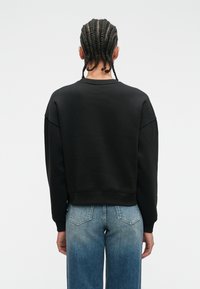 Person with braided hair wearing a black sweatshirt and blue faded jeans, standing facing away against a plain white background.