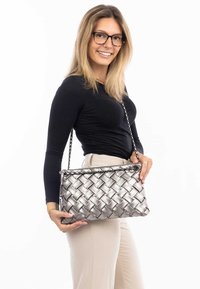 Splotkowana srebrna torebka na łańcuszku, o prostokątnym kształcie i gładkiej teksturze, trzymana przez osobę w czarnej koszuli z długim rękawem.