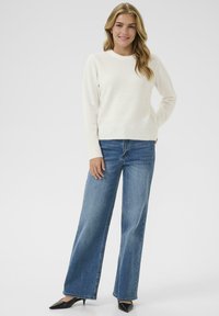 Lichtbeige trui met een ronde hals, gecombineerd met hoge blauwe wijde jeans en zwarte puntige hakken. Moderne casual outfit.
