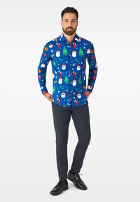 Camicia blu con bottoni caratterizzata da stampe a tema natalizio, tra cui Babbo Natale, alberi e fiocchi di neve; abbinata a pantaloni scuri con fantasia e scarpe nere.