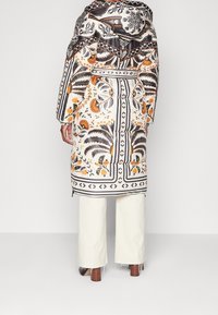 Abrigo acolchado con patrones florales y geométricos en negro, blanco y naranja. Cuenta con una capucha y un cinturón en la cintura. Combinado con pantalones anchos de color crema.