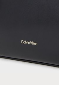 Svart skinnveske med en glatt tekstur, med en gull "Calvin Klein" logo preget på fronten. Firkantet form, minimalistisk design.