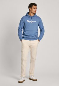 Sudadera azul con bolsillo tipo canguro en la parte frontal y logo blanco, combinada con pantalones cargo beige y zapatillas blancas con suelas marrones.