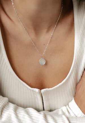 Silver necklace with a round pendant featuring a textured edge, worn on a collarbone above a white ribbed top.