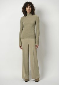 Beige strukturierter Langarmoberteil mit hohem Kragen, kombiniert mit locker sitzenden beige Hosen. Beide Kleidungsstücke haben ein glattes, gewebtes Aussehen.