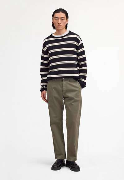 Homme debout, les mains dans les poches, portant une chemise à manches longues rayée noir et blanc, un pantalon vert olive et des chaussures noires sur fond blanc.