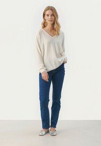 Beiger, langärmeliger V-Ausschnitt-Pullover kombiniert mit dunkelblauen Jeans. Das Model trägt silberne Ballerinas. Einheitlicher Hintergrund.