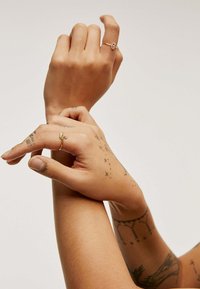 Dos manos con tatuajes, usando un delicado anillo de oro con un diseño en espiral y una simple banda de oro. Textura de piel suave, fondo neutro.