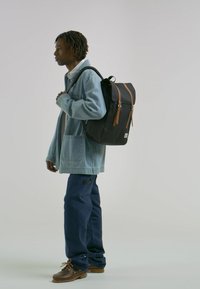 Sac à dos noir avec des accents en cuir marron, porté par une personne vêtue d'une veste en jean bleu clair et d'un pantalon bleu marine, debout devant un fond uni.