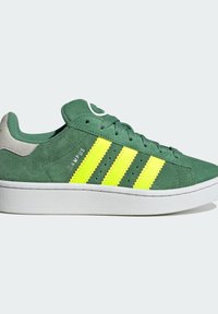 Zapatillas de ante verde con tres franjas amarillas brillantes, suela de goma blanca y acento en el talón gris. Presenta perforaciones a lo largo de los lados.