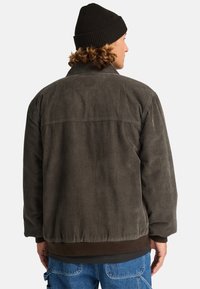 Giacca in corduroy di un verde oliva scuro, caratterizzata da righe verticali, colletto a camicia e una vestibilità rilassata con vita elastica.