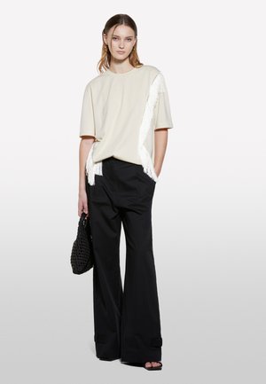 Haut crème à manches courtes avec des franges blanches d'un côté, associé à un pantalon large noir. Sac à main noir texturé à la main.