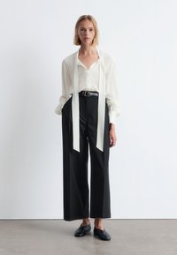 White blouse with long ribbons, black wide-leg trousers, and black slip-on shoes. Smooth fabric and relaxed fit. Simple, monochrome design.