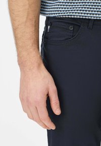 Marineblauwe stoffen broek met een slanke pasvorm, voorzien van een voorzak en contrasterende stiksels. Hand rust op het dijbeen.