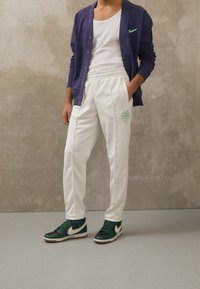 Vita sweatpants med elastisk midja och grön logotyp, parat med en vit linne och lila cardigan, accentuerat av gröna och svarta sneakers.