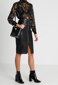 Czarna skórzana ołówkowa spódnica z przednim rozcięciem, zestawiona z prześwitującą, kwiatową bluzką oraz błyszczącymi czarnymi botkami do kostek. Trzyma małą czarną torebkę.