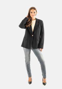 Blazer nero con rever e un solo bottone, abbinato a jeans skinny azzurri e tacchi alti neri. Tessuto morbido, design minimalista.