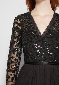 Robe noire à manches longues avec motifs floraux pailletés sur tissu transparent et décolleté en V orné de volants, cintrée à la taille par une bande en velours.