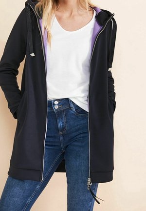 Femme portant une veste à capuche noire zippée avec une doublure violette, une chemise blanche et un jean bleu, les mains dans les poches de la veste.