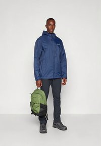 Giacca antipioggia blu navy con cappuccio, materiale leggero, abbinata a pantaloni neri e stivali robusti. Tiene in mano uno zaino verde e nero.