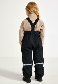 Svarta vattentäta overaller med justerbara remmar, elastisk midja och reflexdetaljer. Bärs över en beige fleece-tröja.