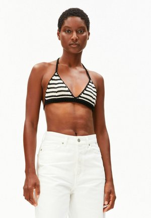 Femme portant un haut de bikini en crochet rayé noir et blanc à col halter et un pantalon blanc taille haute, debout devant un fond uni.