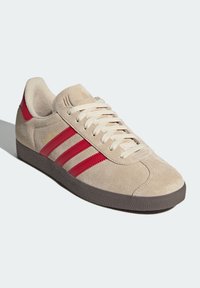 Beige mockask sneaker med röda tre ränder, texturerad sula, vadderad krage och ljusa snörningar. Har minimal branding på sidan.