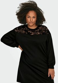 Vestido negro con un panel superior de encaje, mangas largas y un corte holgado. La tela parece suave con una textura visible del diseño de encaje.