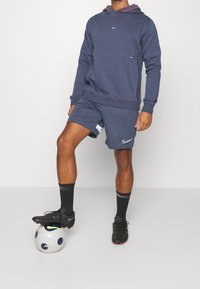 Hoodie Nike bleu marine avec accents rouges, assorti à un short. Le mannequin se tient sur un ballon de football blanc et bleu, portant des chaussures et des chaussettes noires.
