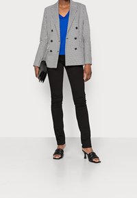 Puku houndstooth na mifumo ya rangi ya buluu na nyeusi, tisheti ya buluu ya v-shingo, suruali za jeans za m_shader muweusi, na viatu vya slay vya m_shader muweusi; ikishikilia clutch ya m_shader muweusi.