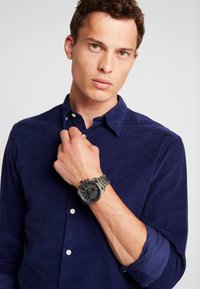 Silver chronograph watch with a black dial, stainless steel band, and three subdials, worn on a hand. Dark blue corduroy button-up shirt.