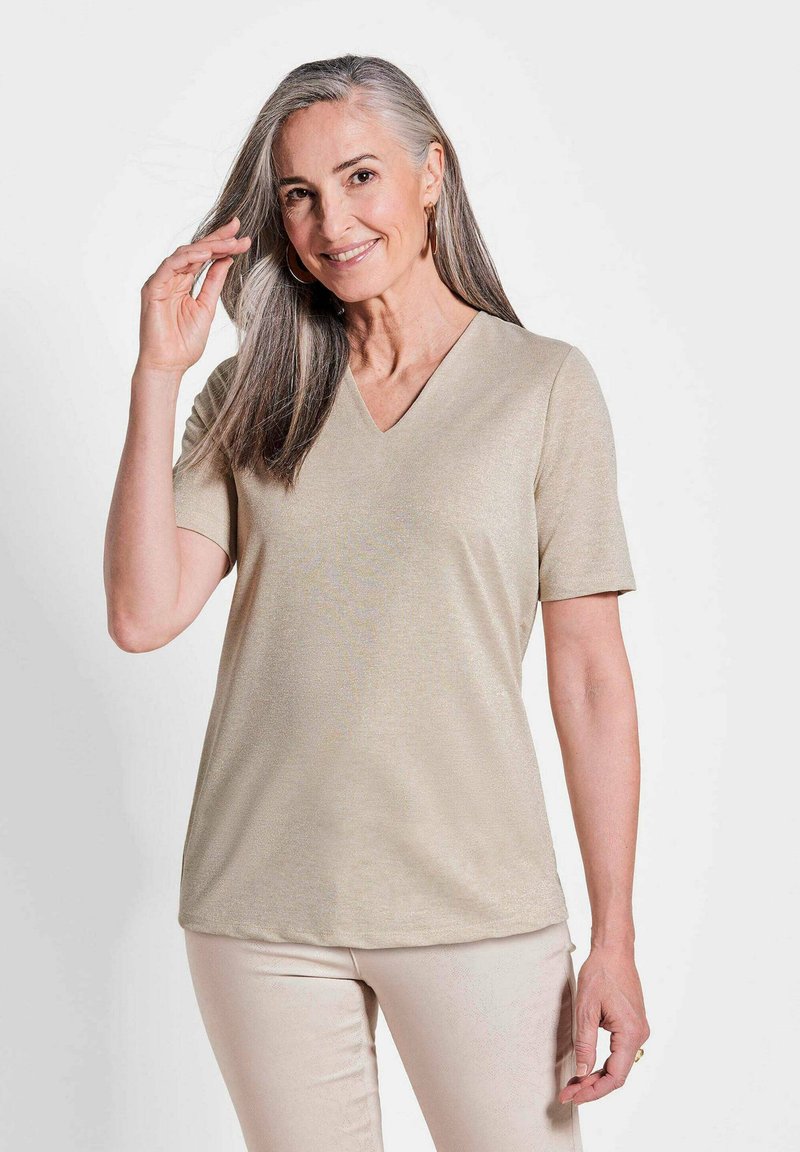 Beige V-hals korte mouwen top van een gladde stof met een subtiele glans, gedragen met lichte broeken; model poseert en glimlacht.