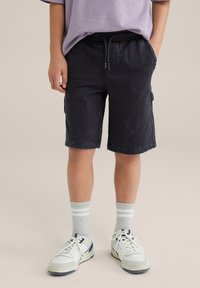 Čierne cargo šortky s pásom na sťahovanie, s vyblednutou textúrou a bočnými vreckami, v kombinácii s bielymi teniskami a šedými pruhovanými ponožkami.