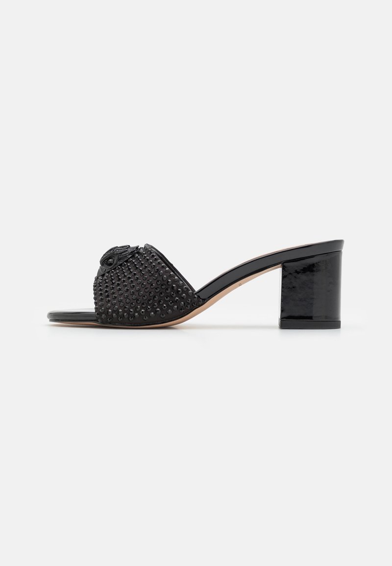 Mule noire à bout ouvert avec un talon bloc moyen, présentant une tige cloutée et un ornement décoratif en forme d’œil à l’avant.