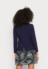 Blazer blu navy con maniche lunghe, dotato di uno spacco posteriore, abbinato a una gonna a strati con stampa floreale scura. Texture liscia e design elegante.