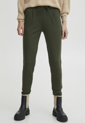 Jogging tapered vert olive avec plis sur le devant et taille ajustable, associés à des bottines noires avec une accent beige.