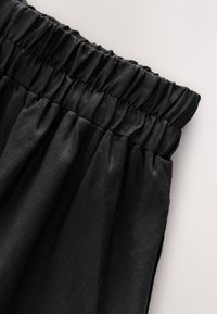 Pantalon en tissu noir avec ceinture élastique froncée et coutures visibles, présenté sur un fond uni clair.